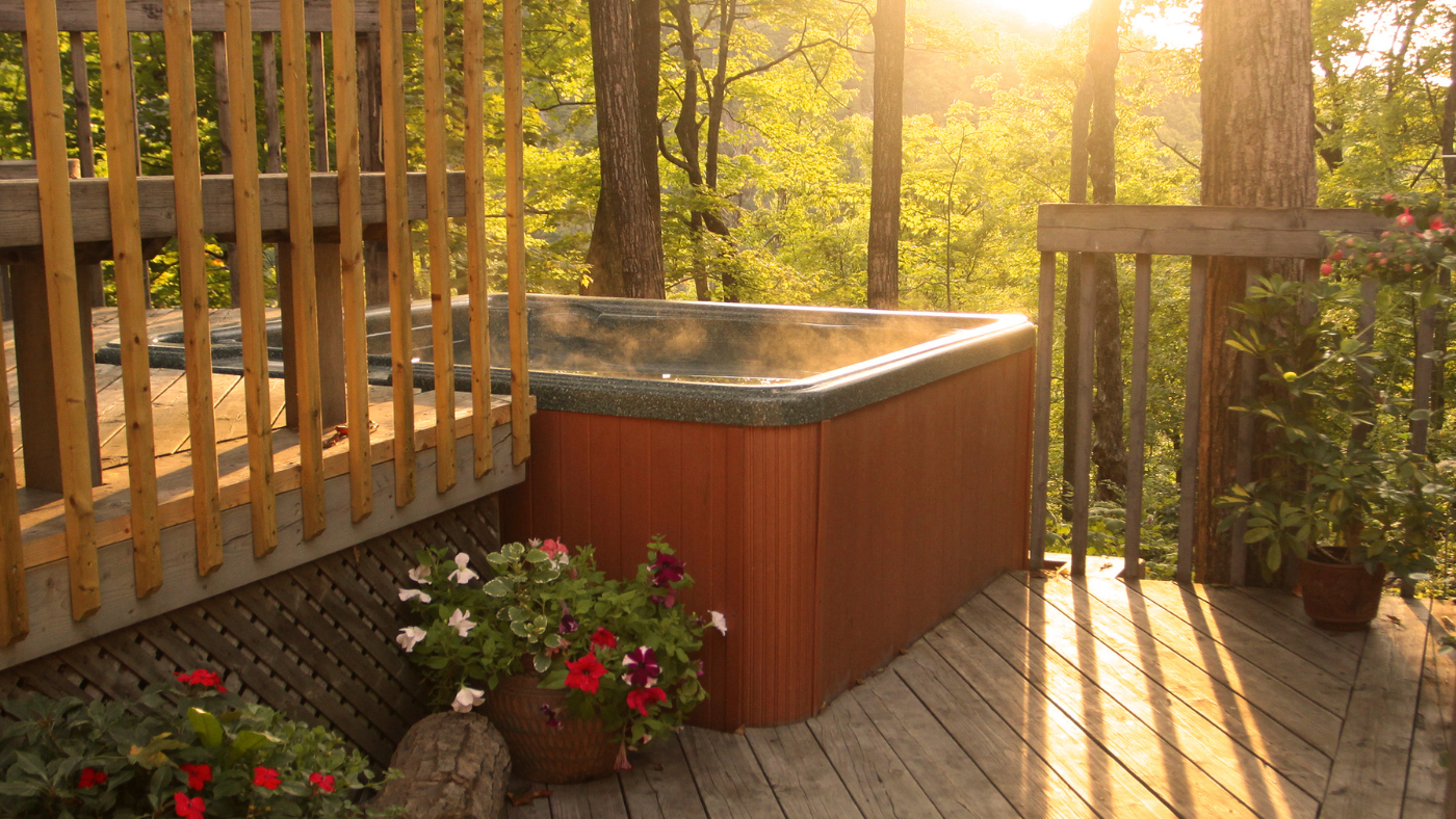 a hot tub with misty steam rising off the water surface sits on a deck in front of a beautiful forest landscape; the sunlight shines through the tree branches and trunks onto the tub and deck floor